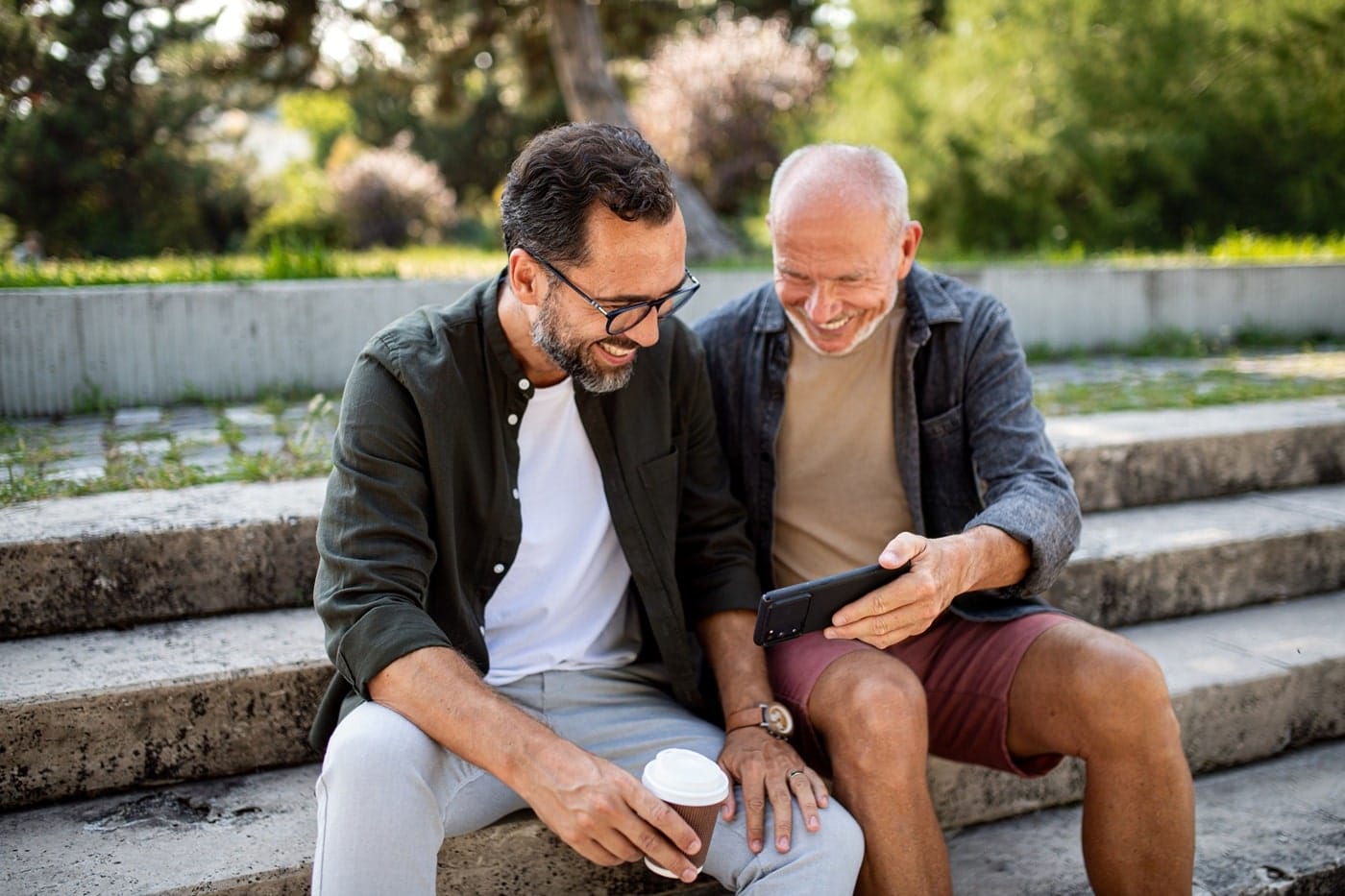 Two men sitting on outdoor steps, one holding a smartphone while they both look at the screen, engaged in conversation.